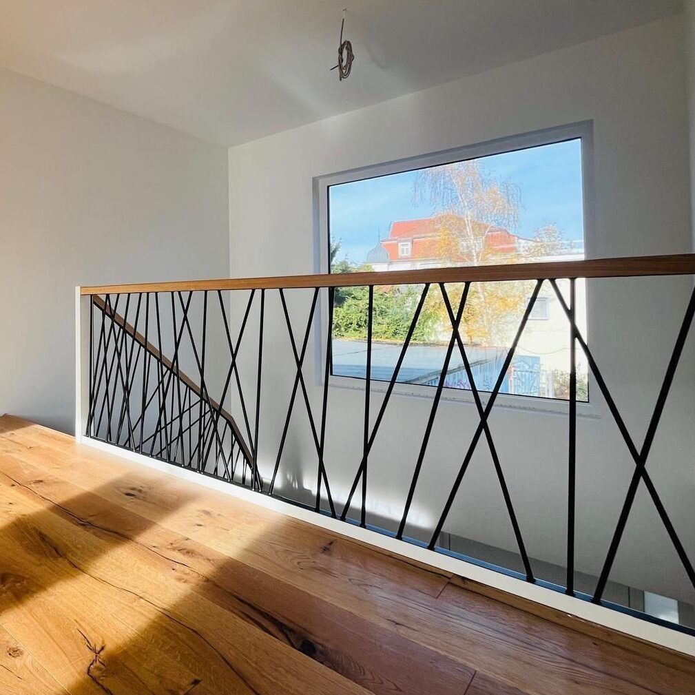Galeriegeländer aus schwarzen, diagonal gekreuzten Metallstäben mit Holzhandlauf vor einem großen Fenster