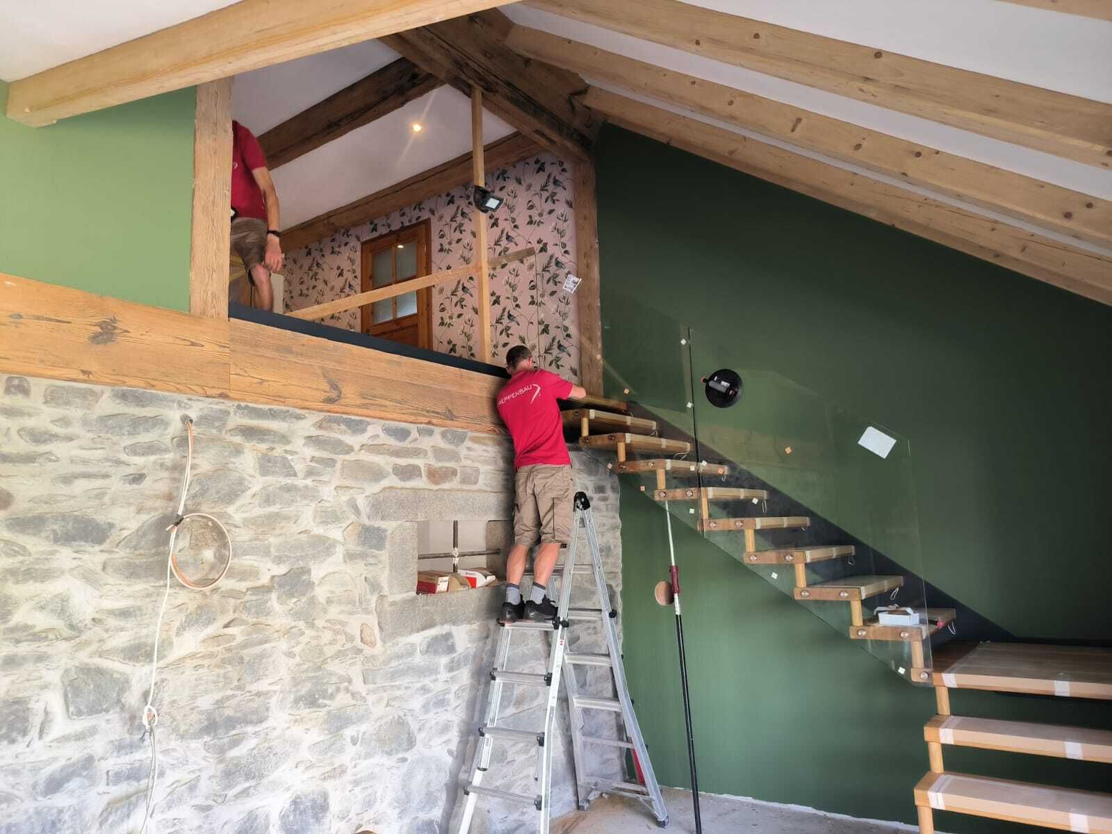 Zwei Monteure bauen eine offene Treppe mit hellen Holzstufen und Glasgeländer an grüner Wand ein