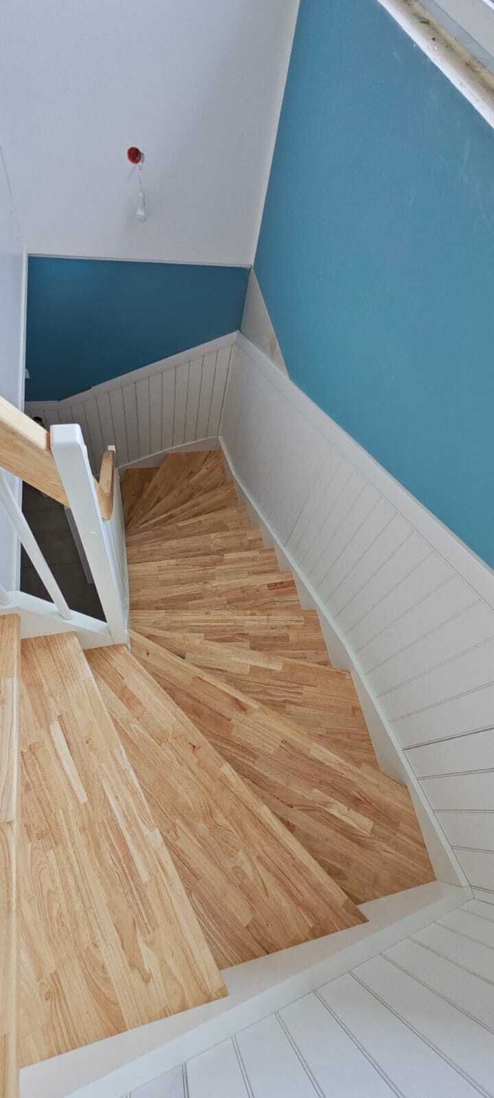 Blick von oben auf eine gewendelte Treppe mit hellen Holzstufen, weißer Paneelverkleidung und blauer Wand