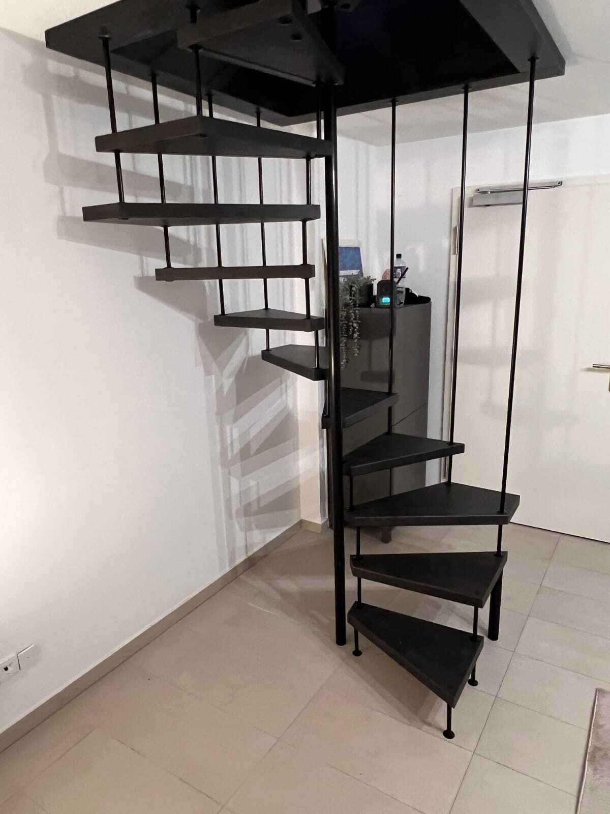 Gewendelte Treppe mit schwarzen Stufen, an senkrechten Metallstäben abgehängt, in einem Flur mit hellem Fliesenboden