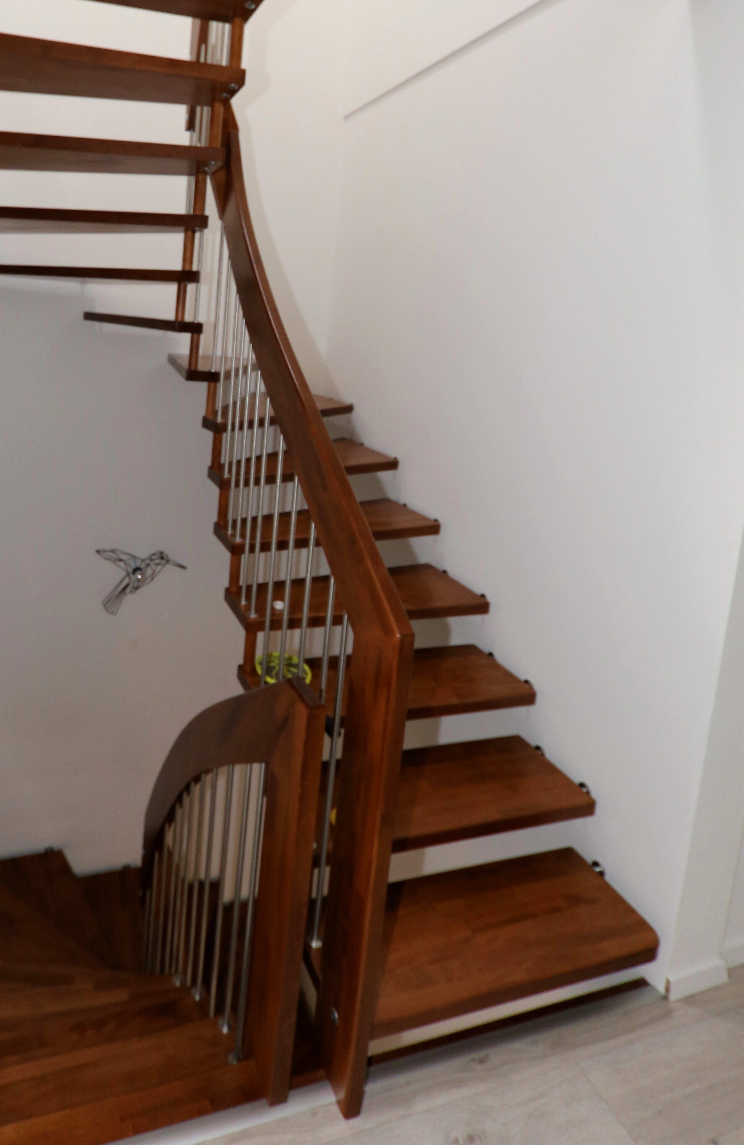 Gewendelte Treppe mit offenen Stufen aus dunklem Holz, Holzhandlauf und senkrechten Edelstahlstäben an weißer Wand