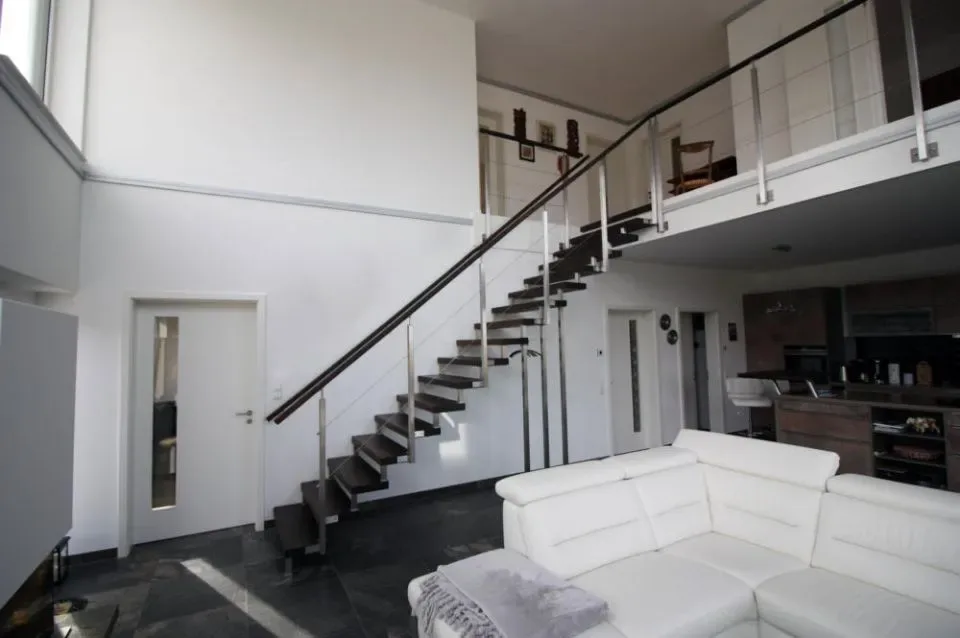 Gerade, offene Treppe mit dunklen Stufen und Edelstahlgeländer führt in einem hohen Wohnraum zur Galerie