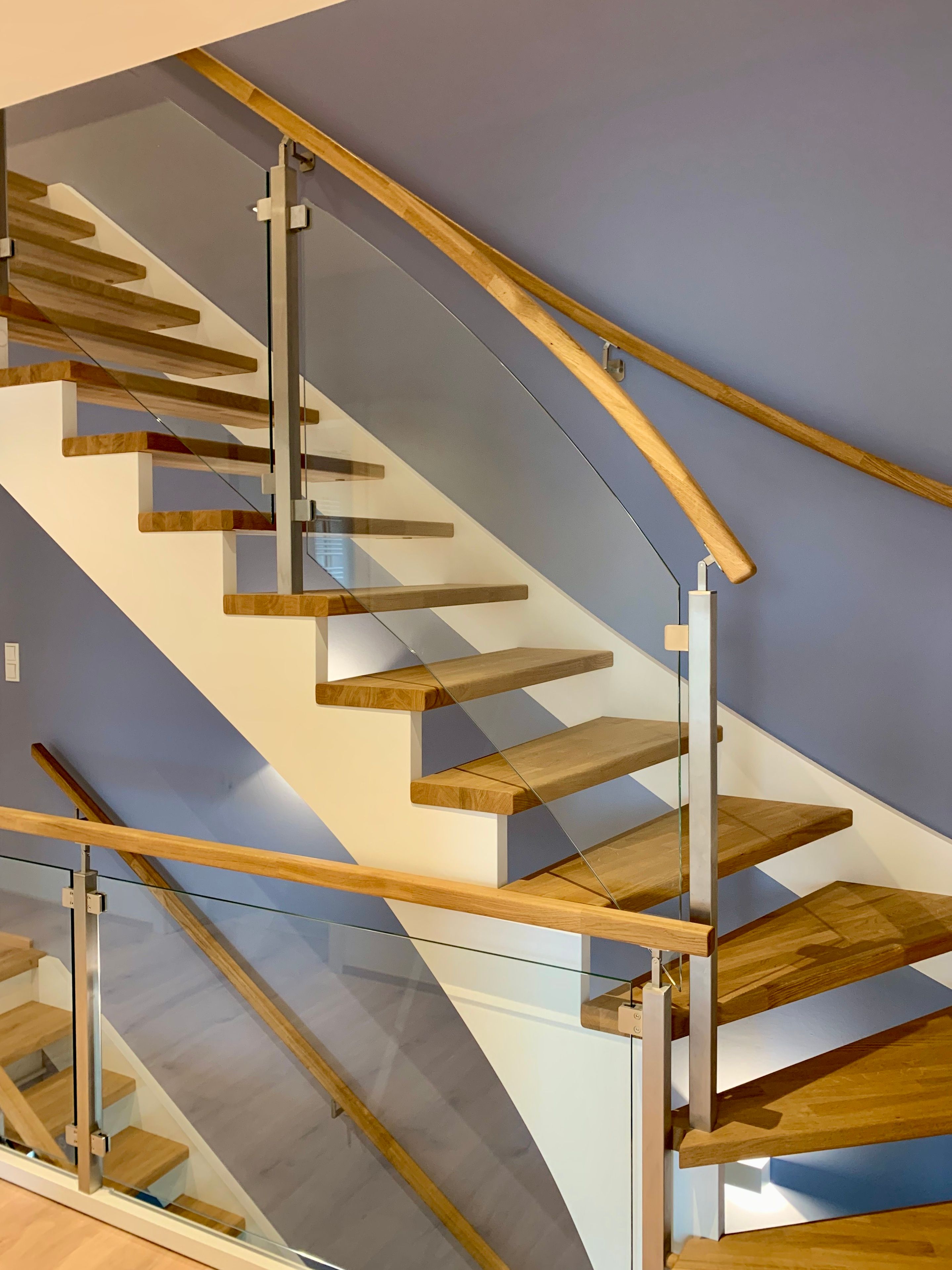 Gewendelte Treppe mit hellen Holzstufen, weißer Wange und Glasgeländer mit Edelstahlpfosten und Holzhandlauf