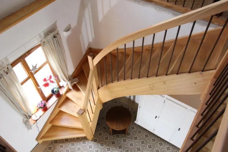 Blick von oben auf eine gewendelte Wangentreppe aus hellem Holz mit schwarzen Metallstäben neben einem Sprossenfenster